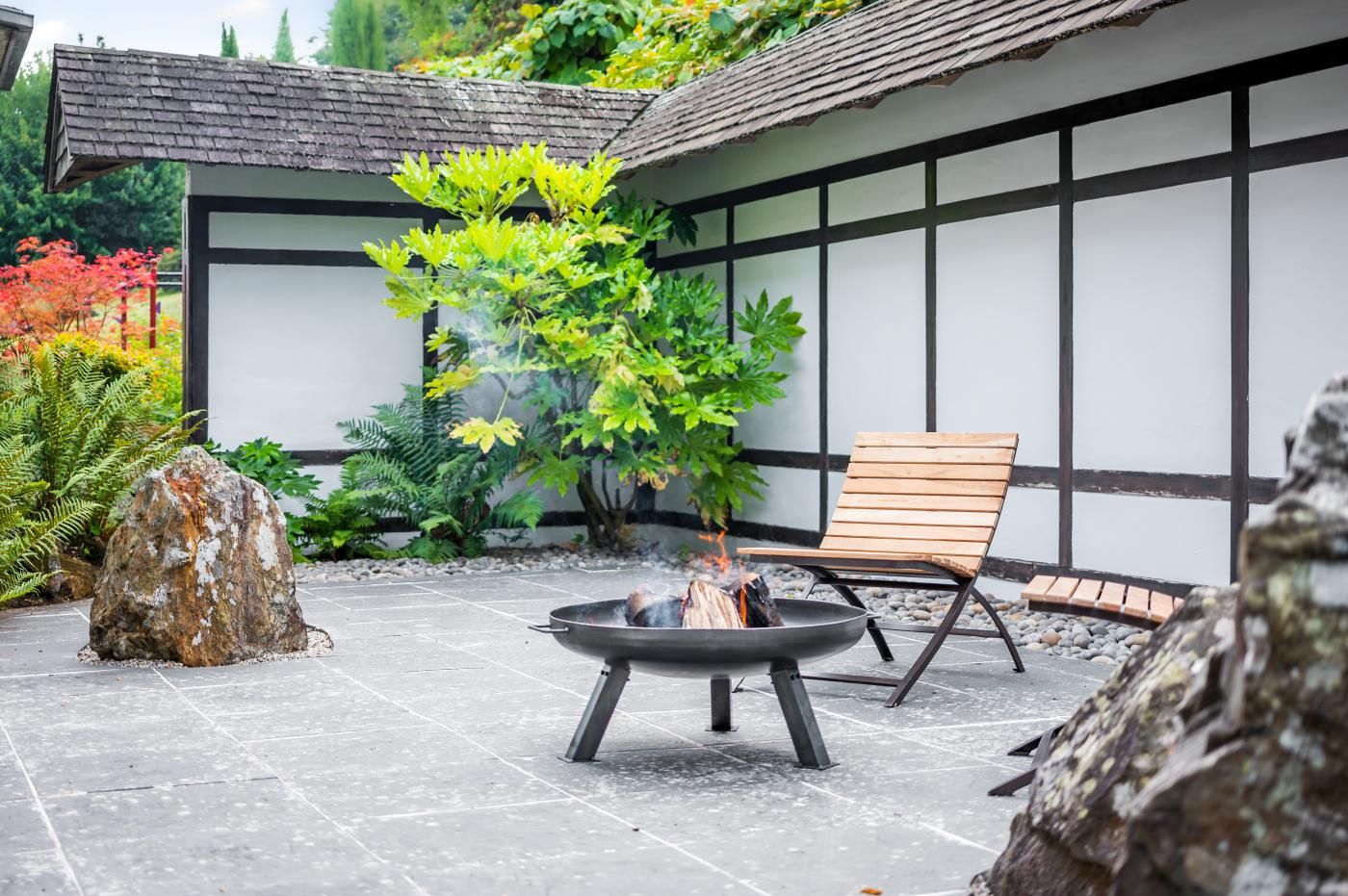 A fire pit with a chair and a large rock in a garden area with a building in the background.