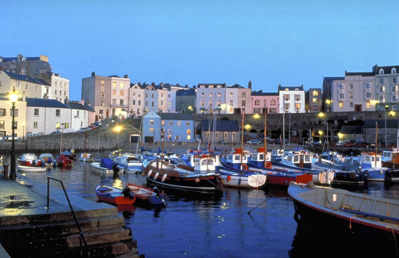 A harbor with boats docked, a stone wall, and buildings in the background.