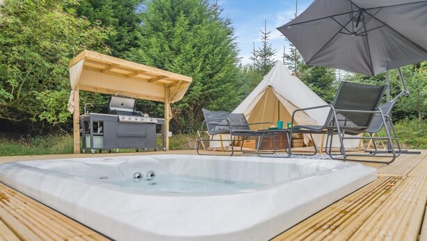 A hot tub with a wooden deck, a grill, and a tent with chairs.