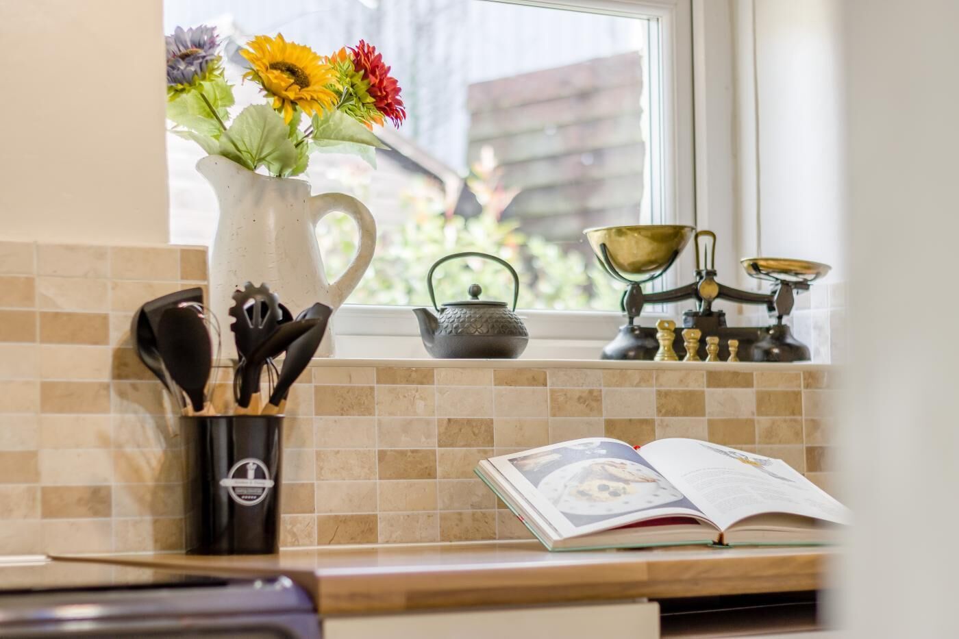 A kitchen counter with a white pitcher, a book, and a window with a view of greenery.