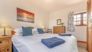 Un letto rifatto con cura, asciugamani blu, un settimino in legno, un comodino con una lampada, un quadro raffigurante un tramonto e una finestra con tende.