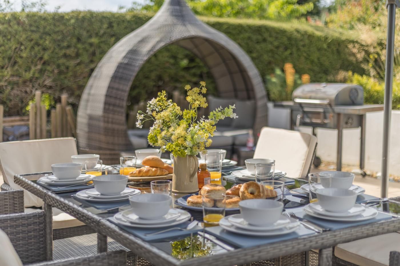 Un espace de restauration en plein air avec une table dressée pour le petit-déjeuner, comprenant des croissants et des viennoiseries, ainsi qu'un vase décoratif rempli de fleurs.