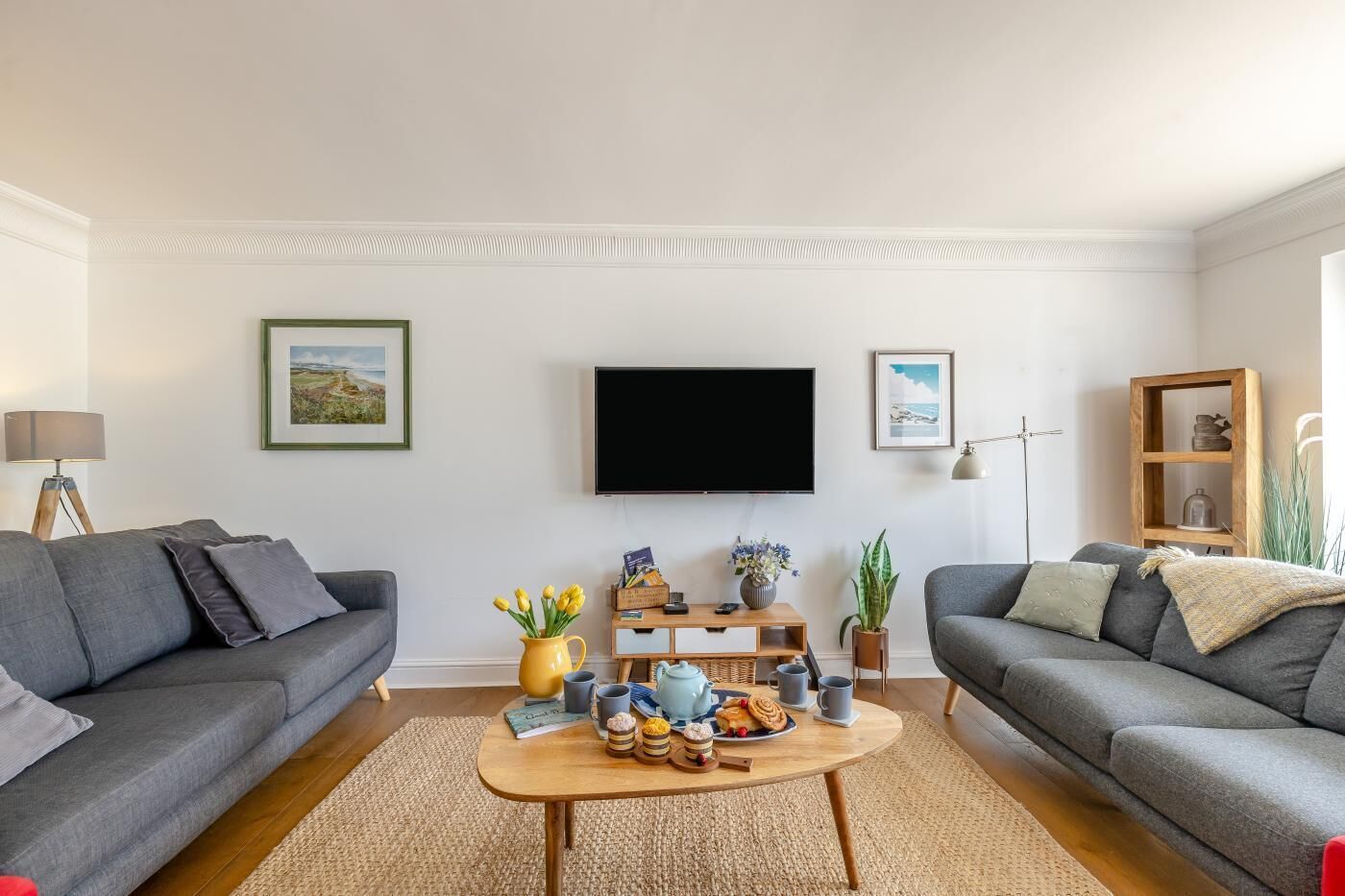 Ein Wohnzimmer mit zwei grauen Sofas, einem Couchtisch mit Snacks, einem Flachbildfernseher und gerahmten Bildern an der Wand.
