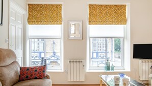 A living room with a sofa, a glass coffee table, two windows with yellow blinds, a radiator, and a television. - 1 Bed in Richmond (oc-k32861) (Richmond)