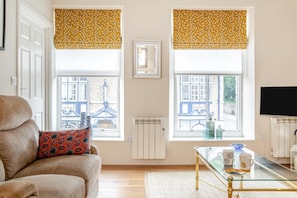 A living room with a sofa, a glass coffee table, two windows with yellow blinds, a radiator, and a television.