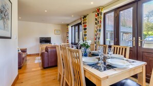 Un comedor con una mesa de madera dispuesta para cuatro personas, rodeada de sillas de madera. Se ve un televisor al fondo y la habitación tiene ventanas amplias con cortinas.