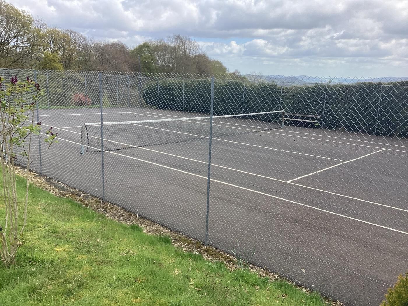 En tennisbana med ett stängsel av sammanlänkade metallkedjor, omgiven av gräs och träd.