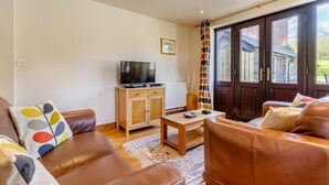 Una sala de estar con sofás de cuero marrón, un mueble de madera con un televisor, una mesita y un ventanal con cortinas.