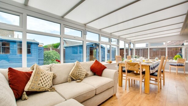 A conservatory with a sofa and dining table, large windows, and a view of a blue house. - 2 Bed in Walcott (oc-2209) (Walcott)