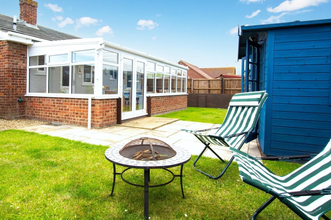 A patio with a fire pit, two striped lounge chairs, a conservatory, and a blue shed.