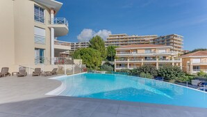 A swimming pool with lounge chairs, a building with balconies, and a clear blue sky. - IMPERIA AP4490 by RIVIERA HOLIDAY HOMES (Niza)