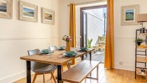 A dining area with a wooden table set for four, two chairs, a vase with flowers, and a view through a glass door to an outdoor area with plants. - 2 Bed in Newquay (oc-j32859) (Newquay)