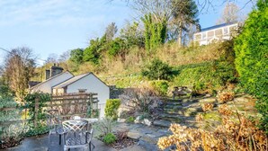 A garden with a stone pathway, a metal table and chairs, and a wooden bench. - 2 Bed in Penrhyndeudraeth (oc-wag442) (Penrhyndeudraeth)