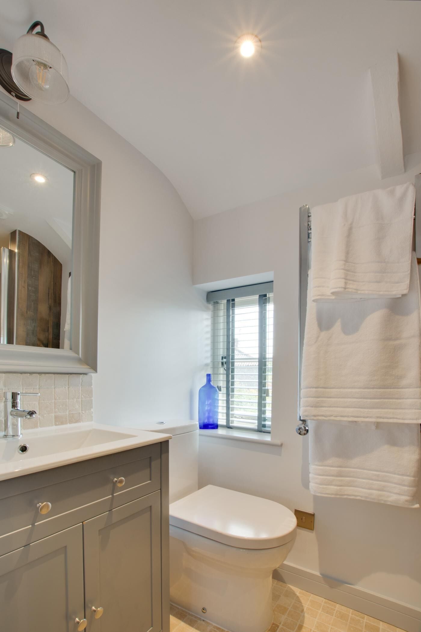 A bathroom with a white sink, toilet, and shower. There is a window with blinds and a towel rack with towels.
