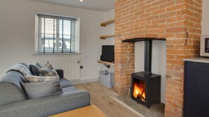 A modern living room with a brick fireplace, a grey sofa, and a television on a wooden shelf. - 1 Bed in Holt (oc-2184) (Holt)