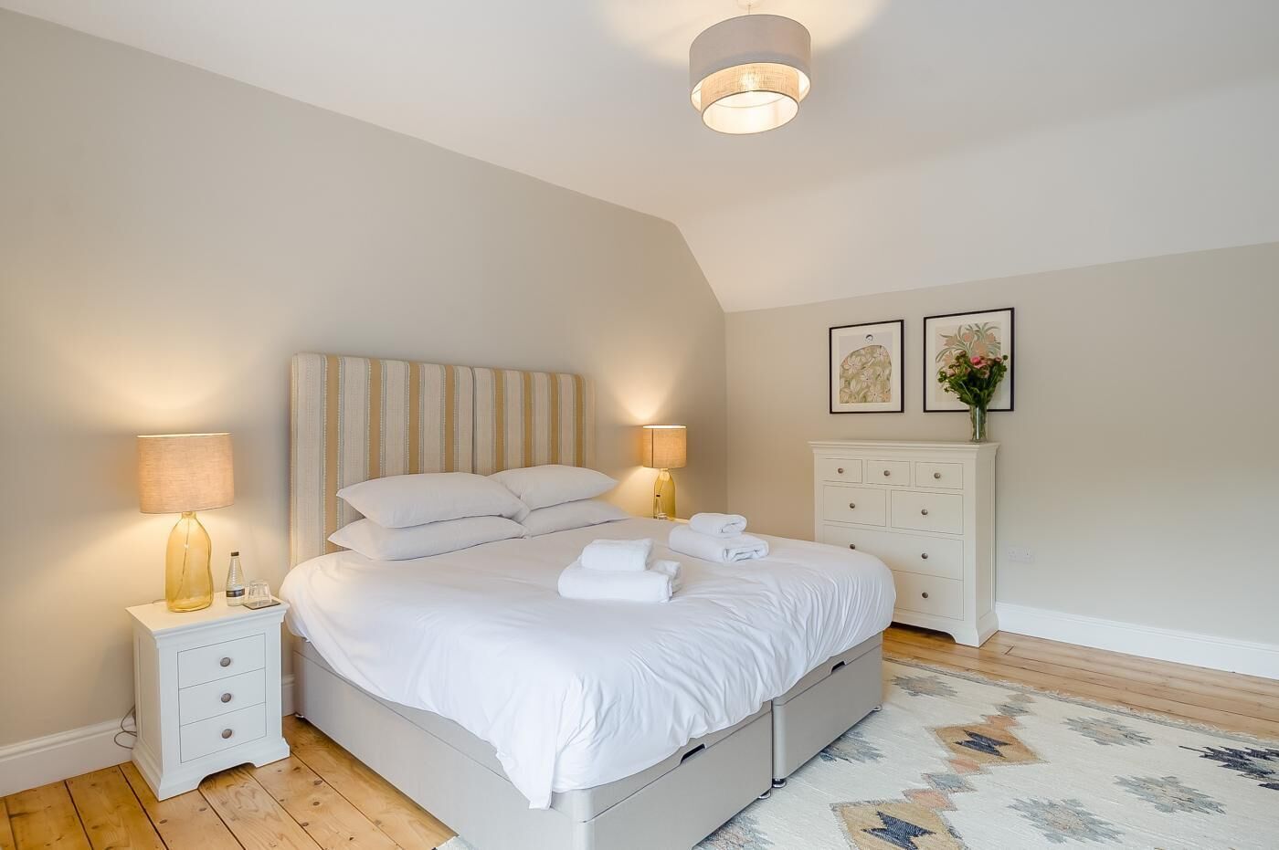 Une chambre à coucher bien aménagée, avec un grand lit, des lampes de chevet, une commode et des œuvres d’art encadrées accrochées au mur.