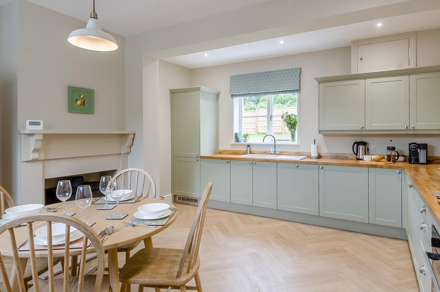A modern kitchen with light grey cabinets, a wooden dining table with chairs, a built-in sink, and a window with a view of the outdoors.