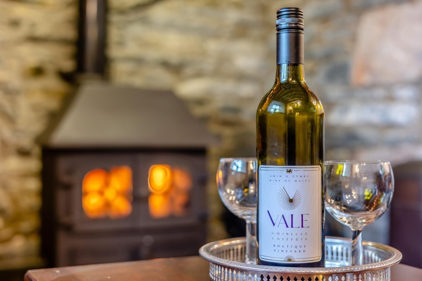 A bottle of wine labeled 'VALE' on a tray with two glasses, set against a stone wall with a fireplace in the background.