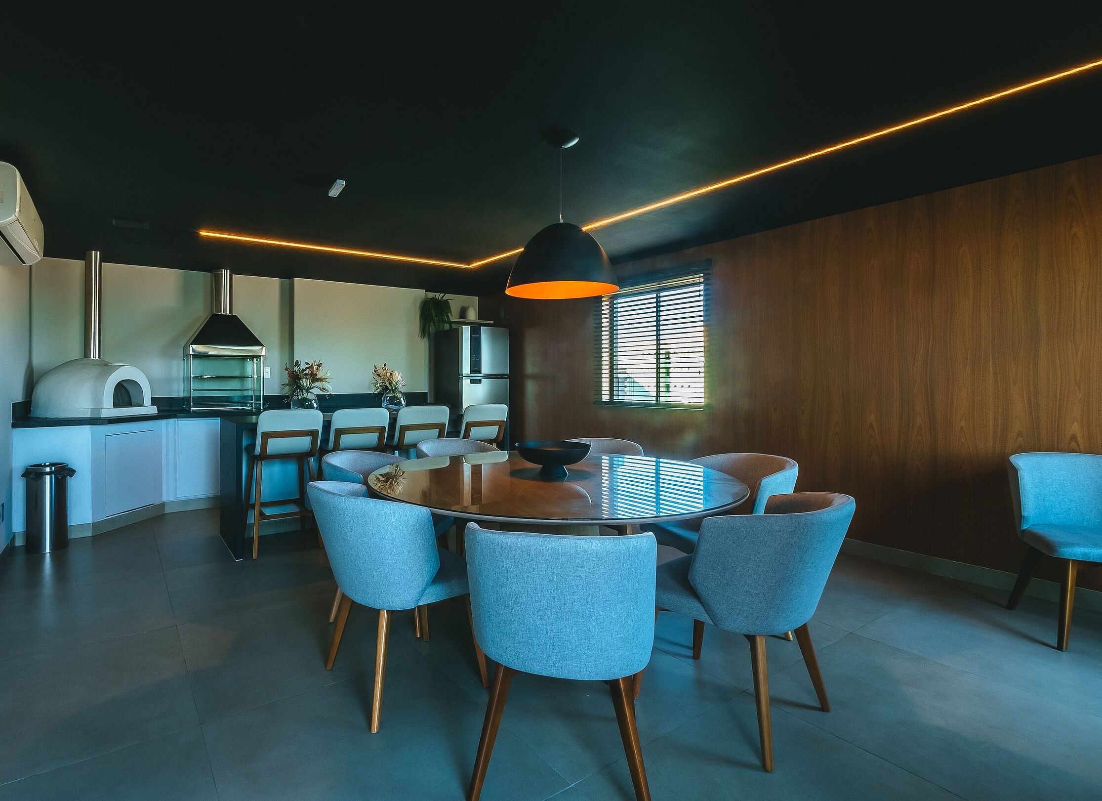 A modern kitchen with a built-in oven, a dining area with a round table and blue chairs, and a large window with blinds.