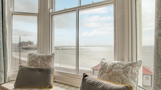 A bay window with two cushions, a glass table with two mugs, and a view of the sea and buildings. - 1 Bed in Aberystwyth (oc-bow286) (Aberystwyth)