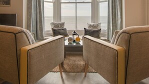 A cozy living room with a large bay window overlooking the sea, featuring two armchairs and a small table with a lamp. - 1 Bed in Aberystwyth (oc-bow286) (Aberystwyth)