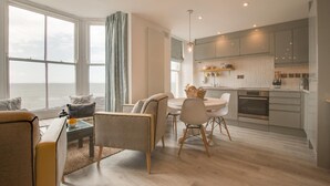 A modern living room with a large window, a dining area, and a kitchen with white cabinets and a built-in oven. - 1 Bed in Aberystwyth (oc-bow286) (Aberystwyth)