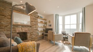 A cozy living room with a stone wall, a large window with a view of the sea, a fireplace, and a modern lamp. - 1 Bed in Aberystwyth (oc-bow286) (Aberystwyth)