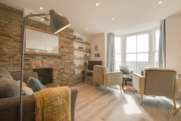 A cozy living room with a stone wall, a large window with a view of the sea, a fireplace, and a modern lamp.
