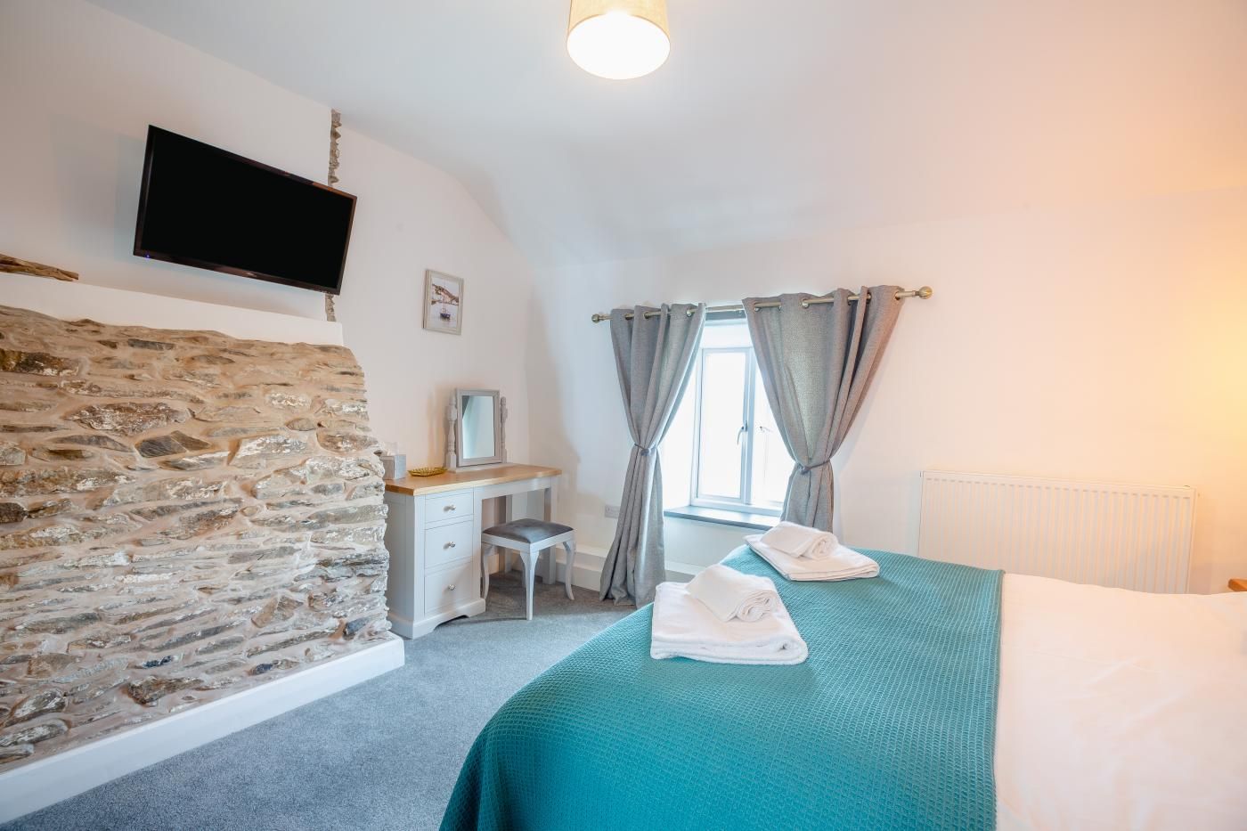 A hotel room with a stone wall, a flat-screen TV, a desk with a chair, a bed with a teal bedspread, and a window with curtains.