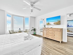 A spacious bedroom with a large bed, a ceiling fan, a wooden dresser, and a beach-themed painting on the wall. - 🏝️Beach FRONT | 2 POOLS | Hot Tub | Emerald Beach 931🏝️ (Panama City Beach)