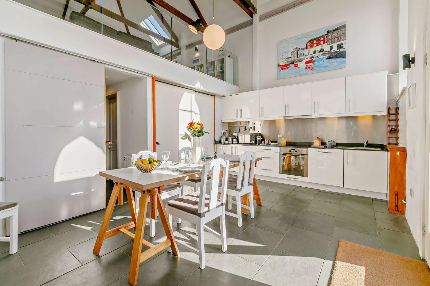 Une cuisine moderne avec des meubles blancs, un coin repas avec une table et des chaises en bois, et un tableau représentant des bateaux accroché au mur.
