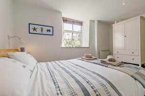 A bedroom with a bed, a nightstand, a lamp, a window with blinds, a wardrobe, and a framed picture on the wall. - 2 Bed in Wadebridge (oc-p00449) (Wadebridge)