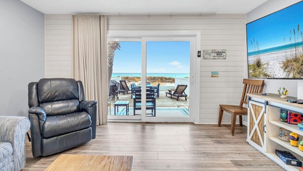 A living room with a leather chair, a wooden coffee table, and a TV on a wooden shelf. There is a sliding glass door leading to a patio with outdoor furniture and a view of the beach. - 🌊Beachfront | Dog Friendly | Incredible Gulf Views | Surfs Up🌊 (Panama City)