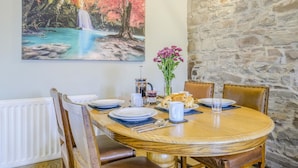 A dining table set with plates, cutlery, and a vase of flowers, surrounded by chairs in a room with a stone wall and a painting of a waterfall. - 2 Bed in Cray (oc-bow287) (Crai)