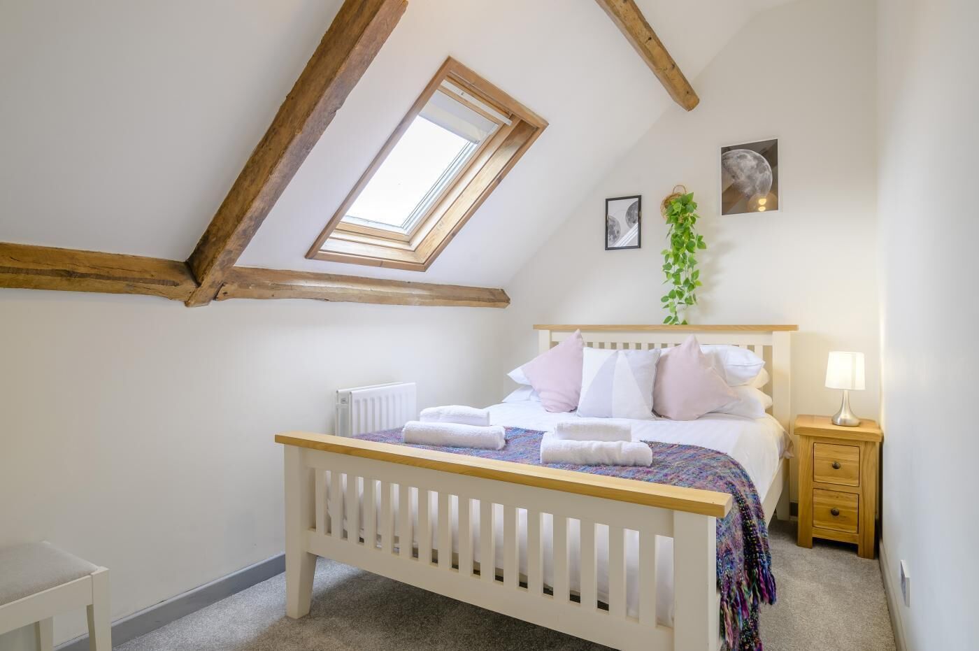 A bedroom with a sloped ceiling, a skylight, a bed with white bedding, a bedside table with a lamp, and a radiator.