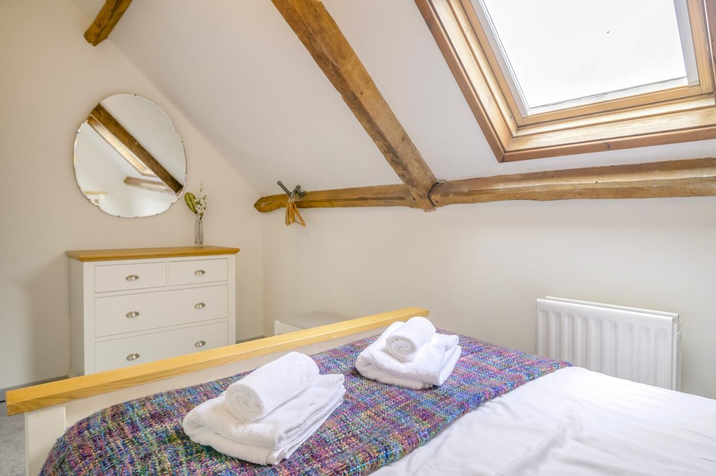 Une chambre avec un plafond en pente, une tête de lit en bois, une commode blanche, un miroir, un radiateur et un lit recouvert d’une couverture colorée.