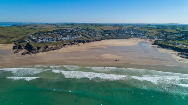 Vue aérienne d’une ville côtière avec une plage de sable, des eaux d’un bleu limpide et un quartier résidentiel avec des maisons.