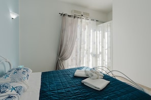 A neatly made bed with a blue quilt, a window with sheer curtains, and a wall-mounted air conditioner.