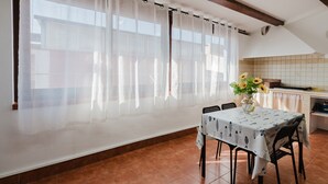 Ein Essbereich mit Tisch für zwei Personen, einem Fenster mit durchsichtigen Vorhängen und einer Küche im Hintergrund.