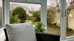 A room with a large window, a wicker chair with a white cushion, and a view of a garden with a house in the background. - 3 Bed in Winterton-on-Sea (oc-2205) (Winterton-on-Sea)