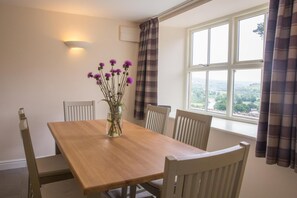A dining area with a wooden table and four chairs, a vase with purple flowers, a wall-mounted light, and a window with a view of greenery. - 3 Bed in Low Row (oc-ds1030) (Low Row)
