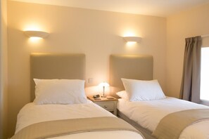 A hotel room with two beds, each with white bedding and headboards. There is a nightstand with a lamp and a phone between the beds. - 3 Bed in Low Row (oc-ds1030) (Low Row)