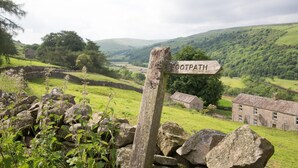 A wooden signpost labeled "FOOTPATH" in a rural landscape with stone walls, greenery, and traditional buildings. - 3 Bed in Low Row (oc-ds1030) (Low Row)