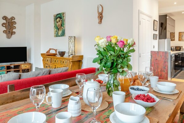 Un coin repas avec une table en bois mise pour le petit-déjeuner, un canapé à l’arrière-plan et un téléviseur sur le côté gauche.