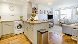 A modern kitchen with a washing machine, a dining area with a table and chairs, a living room with a sofa, and a TV. - 2 Bed in Cromer (oc-2202) (Cromer)