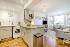 A modern kitchen with a washing machine, a dining area with a table and chairs, a living room with a sofa, and a TV. - 2 Bed in Cromer (oc-2202) (Cromer)