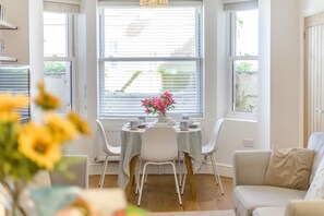 A dining area with a table set for two, a window with blinds, and a view of greenery outside. - 2 Bed in Cromer (oc-2202) (Cromer)
