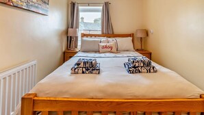 A bedroom with a wooden bed, two bedside tables with lamps, a window with curtains, and a cityscape painting on the wall. - 1 Bed in Whitby (oc-w121) (Whitby)