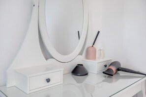 A white vanity with a large mirror, a black hairdryer, a pink vase, and a small container.