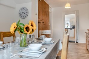 Comedor con mesa de madera dispuesta para cuatro, un reloj en la pared y una cocina visible al fondo.